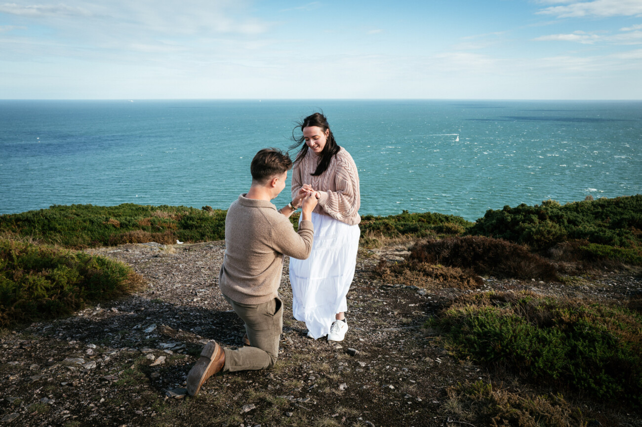 A Secret Proposal at Howth Cliffs & a Private Hour at Howth Castle: Joseph & Shannon's Engagement Shoot 3 A person kneels and holds hands with another person on a rocky cliff overlooking the sea under a partly cloudy sky.