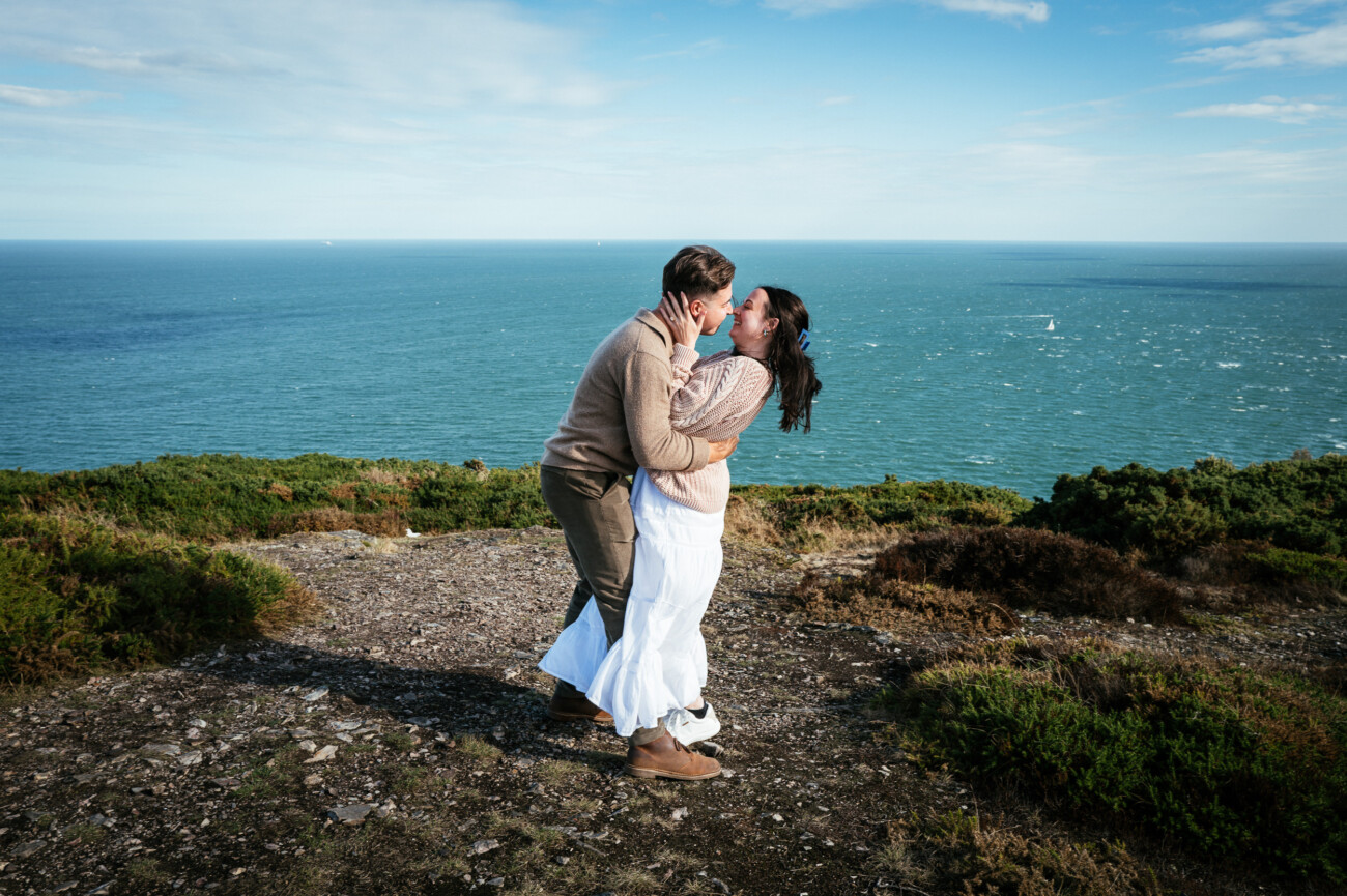 A Secret Proposal at Howth Cliffs & a Private Hour at Howth Castle: Joseph & Shannon's Engagement Shoot 4 A couple embraces and smiles on a rocky cliff overlooking the sea under a partly cloudy sky.