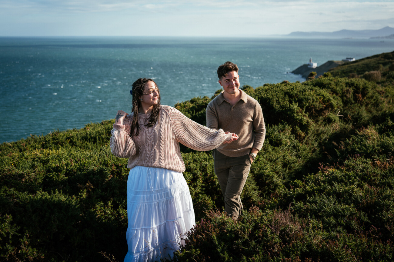A Secret Proposal at Howth Cliffs & a Private Hour at Howth Castle: Joseph & Shannon's Engagement Shoot 5 Two people walk along a green, coastal hillside with the sea and distant mountains in the background under a partly cloudy sky.