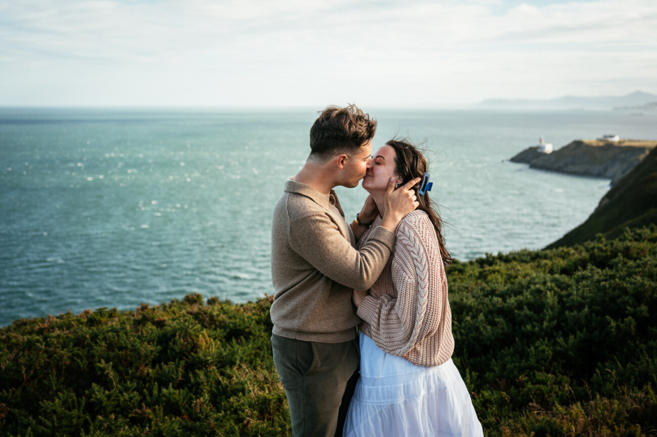 A Secret Proposal at Howth Cliffs & a Private Hour at Howth Castle: Joseph & Shannon's Engagement Shoot 6 A couple snogs on a grassy cliff overlooking the sea, with a lighthouse visible in the background under a partly cloudy sky.