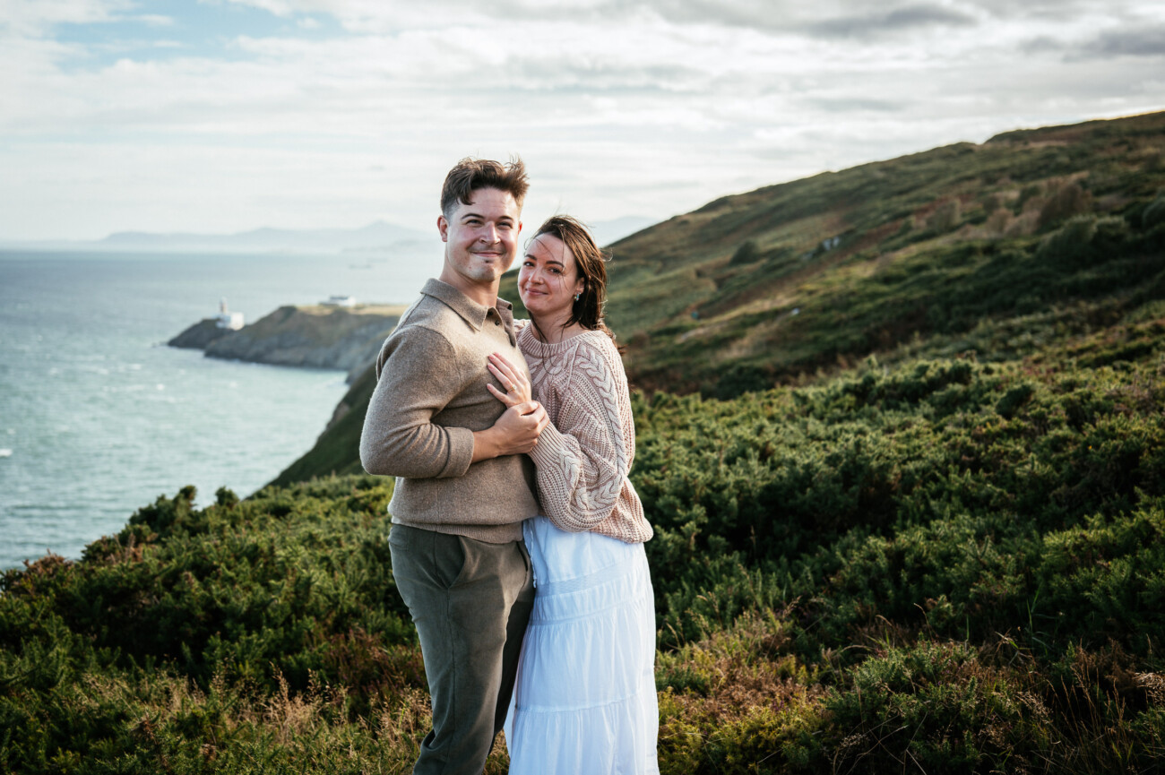 A Secret Proposal at Howth Cliffs & a Private Hour at Howth Castle: Joseph & Shannon's Engagement Shoot 7 A couple stand close together on a grassy hillside with the sea, cliffs, and a lighthouse visible in the background under a partly cloudy sky.