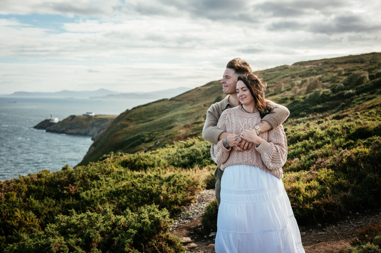 A Secret Proposal at Howth Cliffs & a Private Hour at Howth Castle: Joseph & Shannon's Engagement Shoot 8 A couple stands on a grassy cliffside overlooking the sea, with the man hugging the woman from behind as they both look content.