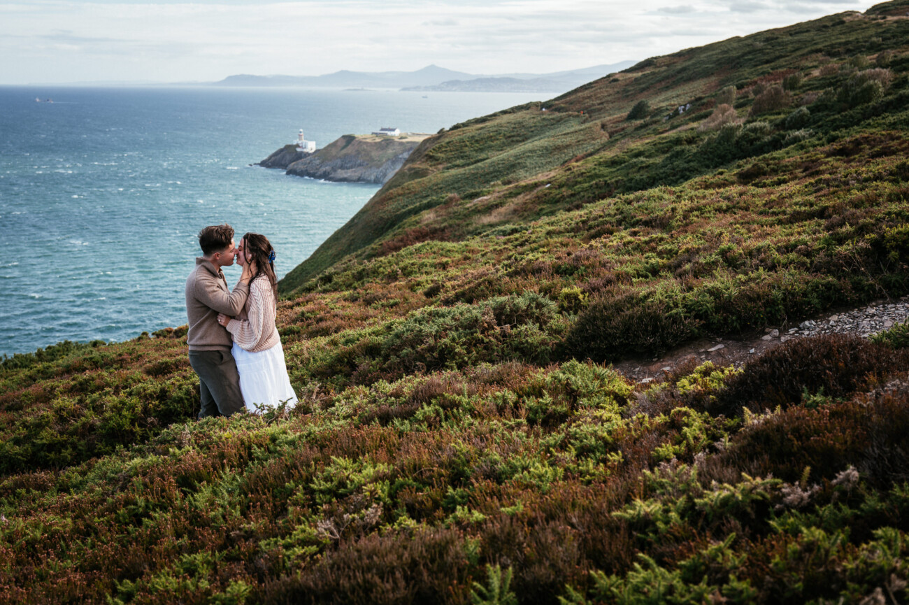 A Secret Proposal at Howth Cliffs & a Private Hour at Howth Castle: Joseph & Shannon's Engagement Shoot 9 A couple stand close together on a grassy hillside overlooking the sea, with a lighthouse visible in the distance under a cloudy sky.