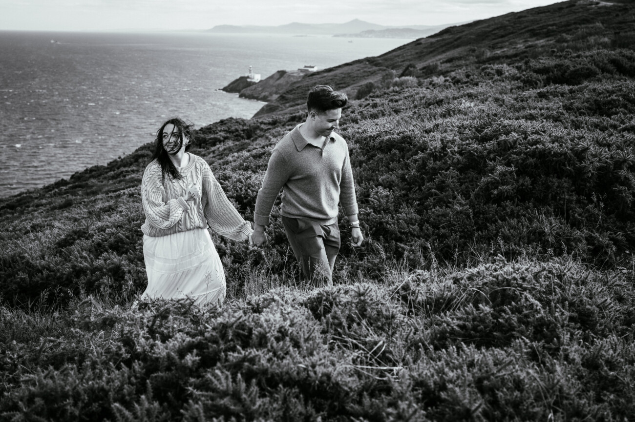 A Secret Proposal at Howth Cliffs & a Private Hour at Howth Castle: Joseph & Shannon's Engagement Shoot 10 A couple walks hand in hand up a grassy hillside overlooking the sea, with distant hills and a lighthouse visible under a cloudy sky.