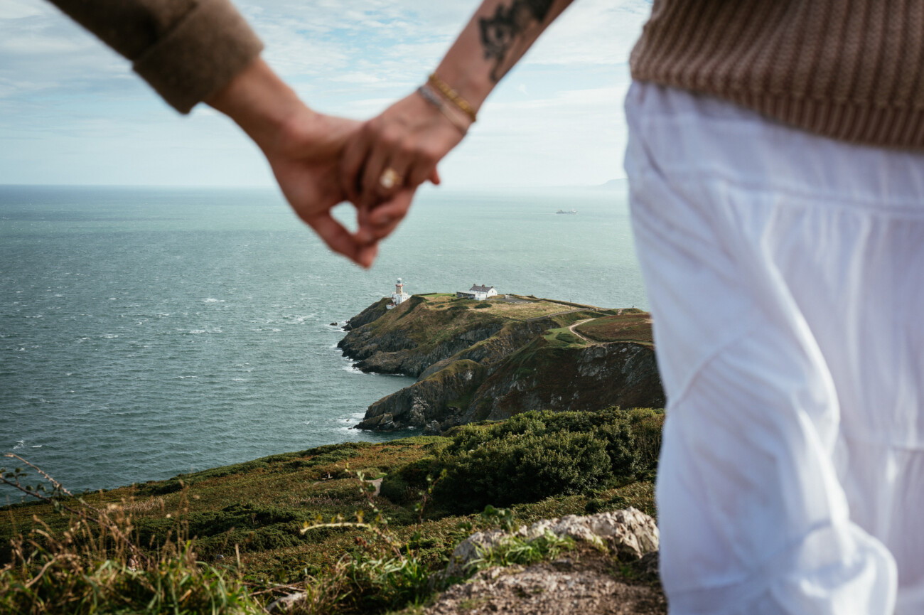 A Secret Proposal at Howth Cliffs & a Private Hour at Howth Castle: Joseph & Shannon's Engagement Shoot 11 Two people hold hands in the foreground, overlooking a coastal cliff with a lighthouse and buildings by the sea in the distance.