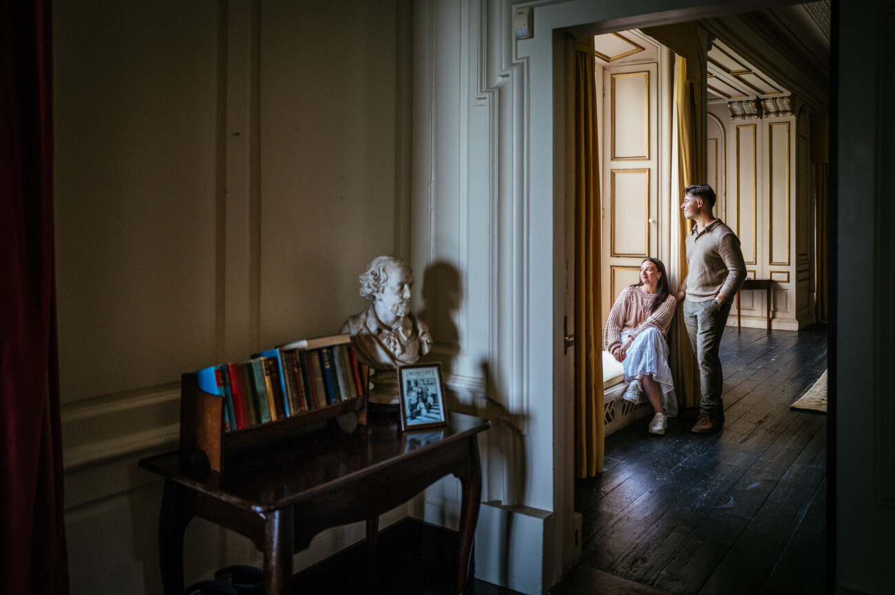 A Secret Proposal at Howth Cliffs & a Private Hour at Howth Castle: Joseph & Shannon's Engagement Shoot 12 A man and woman stand by a window in a sunlit room; nearby, a table holds books, a framed photo, and a bust during their Howth Castle private engagement photoshoot.