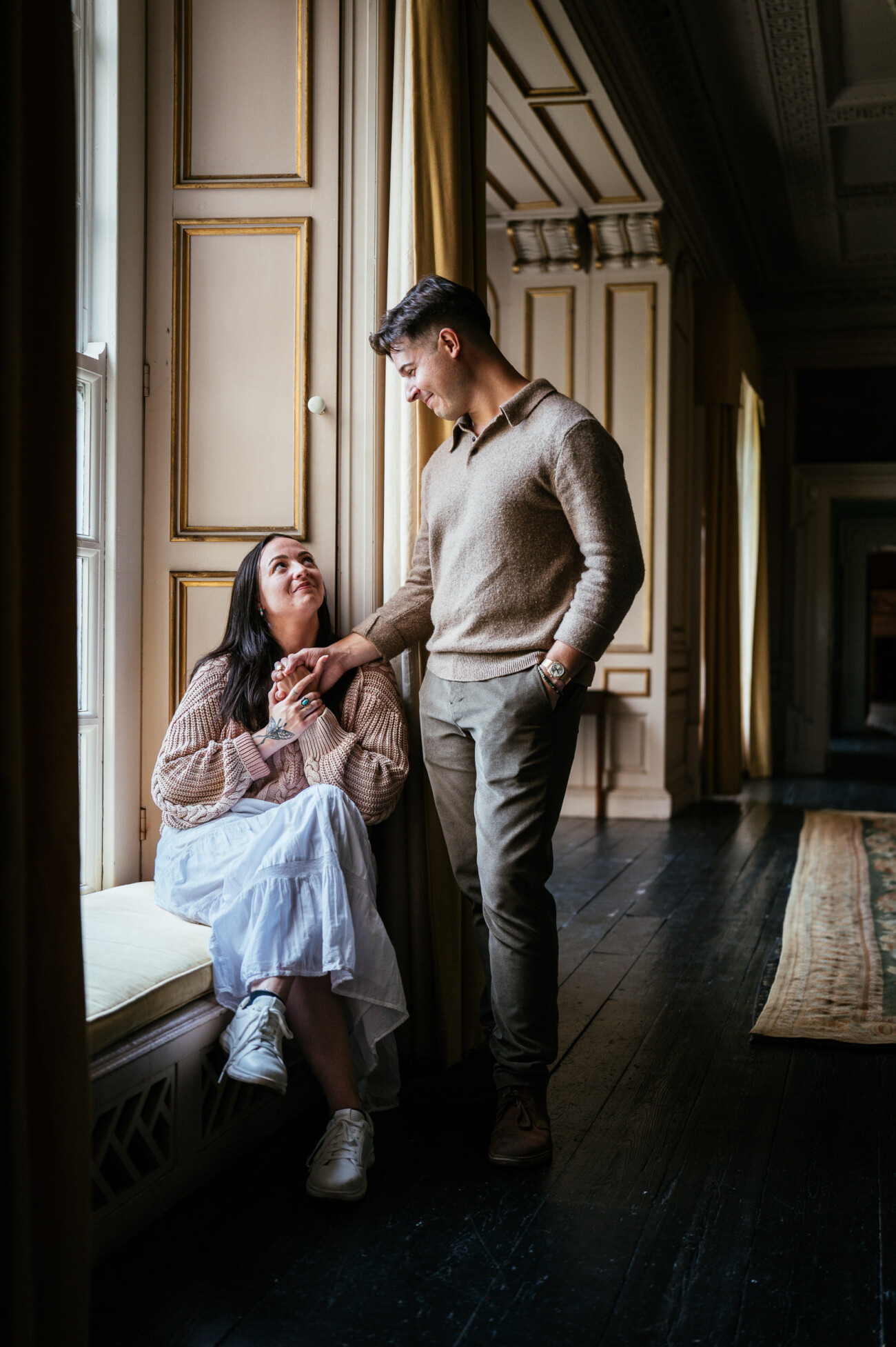 A Secret Proposal at Howth Cliffs & a Private Hour at Howth Castle: Joseph & Shannon's Engagement Shoot 13 A woman sits by a window holding a man’s hand as he stands beside her in a sunlit, ornate corridor with wooden floors.
