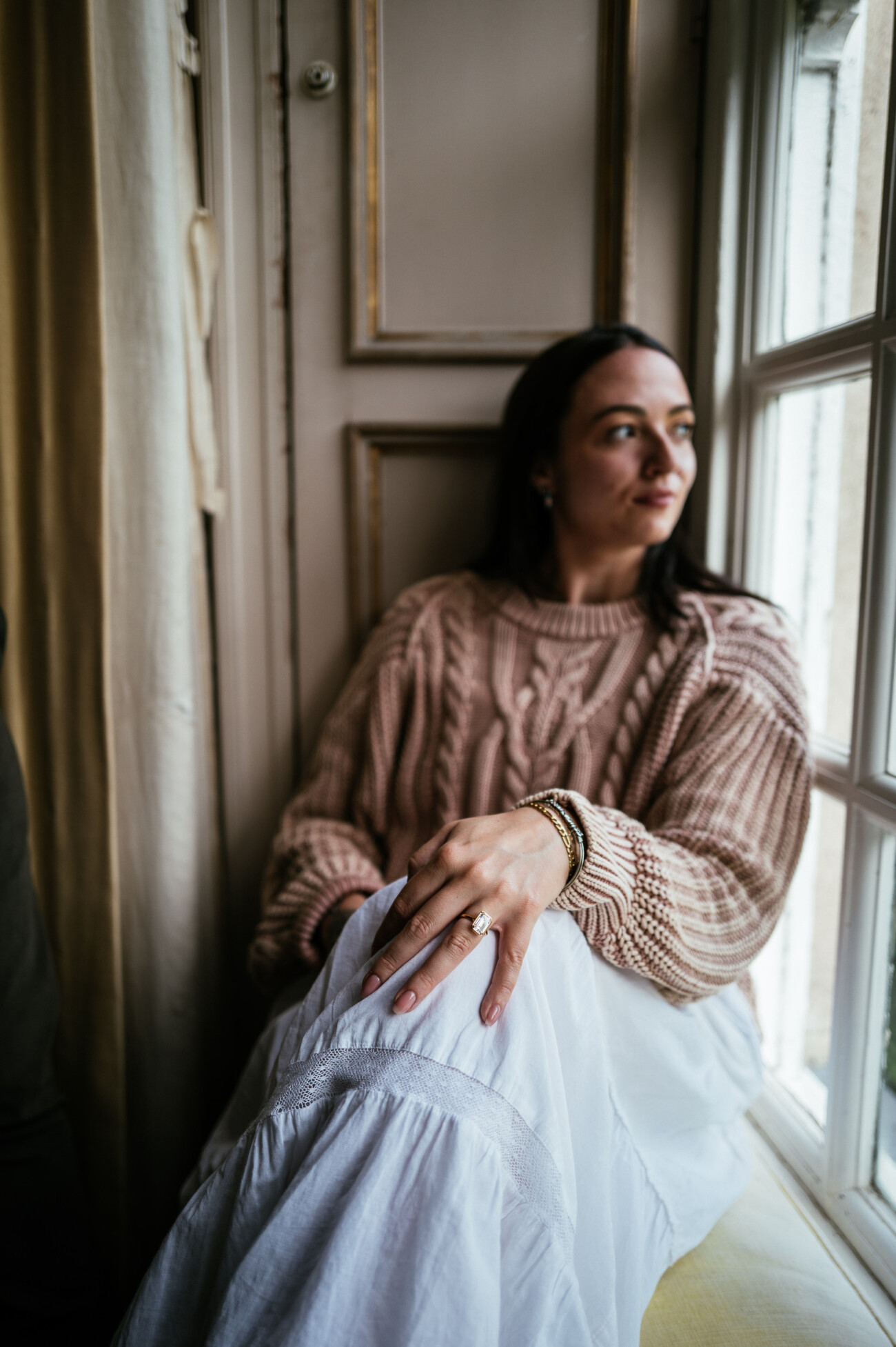A Secret Proposal at Howth Cliffs & a Private Hour at Howth Castle: Joseph & Shannon's Engagement Shoot 14 Woman in a beige jumper and white skirt sits by a window, looking outside with a thoughtful expression, her hand resting on her knee.