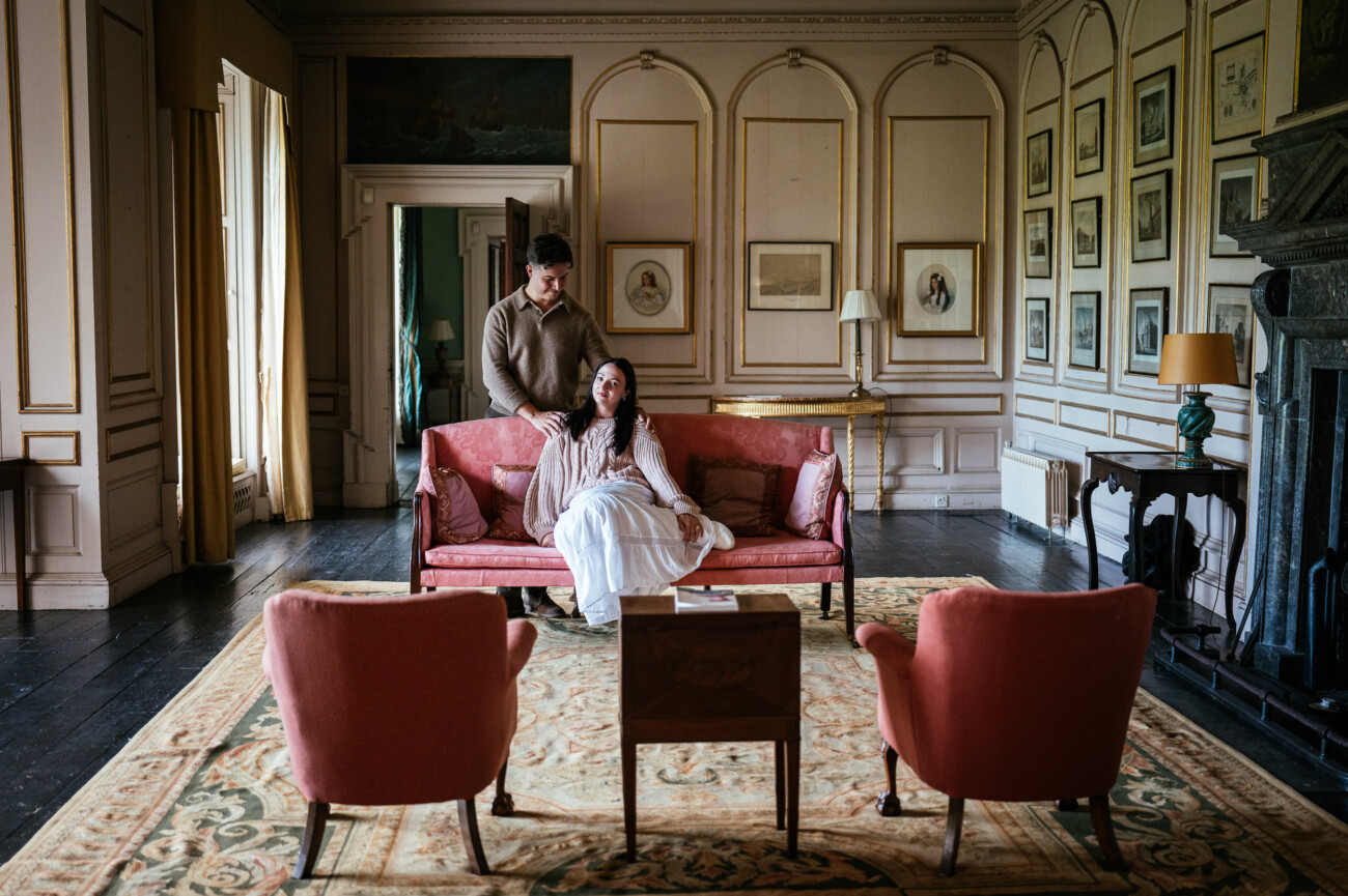 A Secret Proposal at Howth Cliffs & a Private Hour at Howth Castle: Joseph & Shannon's Engagement Shoot 15 A man stands behind a seated woman on a pink sofa in a vintage-style living room with framed art and pink chairs.
