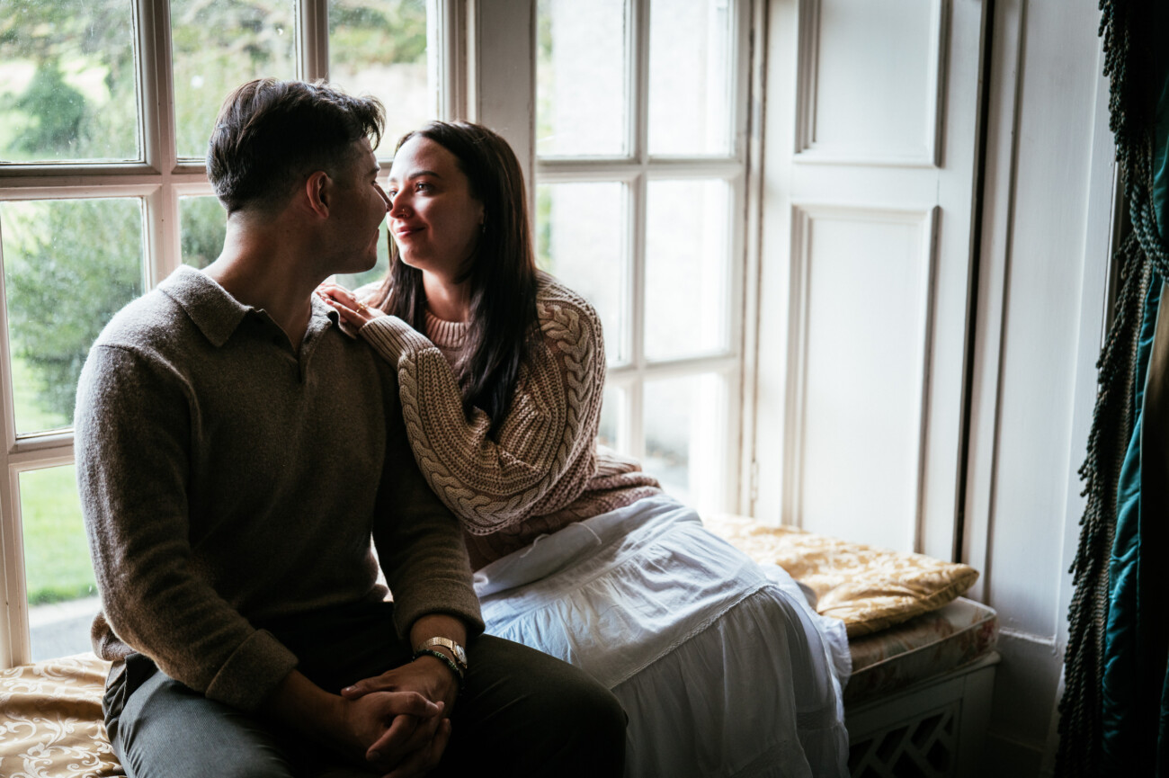 A Secret Proposal at Howth Cliffs & a Private Hour at Howth Castle: Joseph & Shannon's Engagement Shoot 16 A man and woman sit close together on a window seat, facing each other and smiling, with natural light coming through the large windows.
