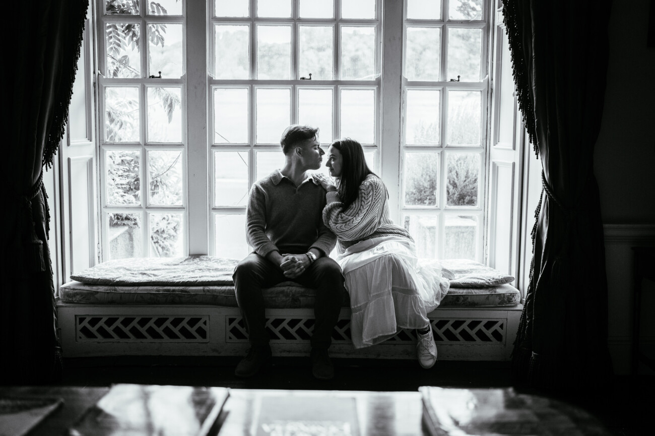 A Secret Proposal at Howth Cliffs & a Private Hour at Howth Castle: Joseph & Shannon's Engagement Shoot 17 A couple sits closely together on a window seat, holding hands and looking at each other in a softly lit room.