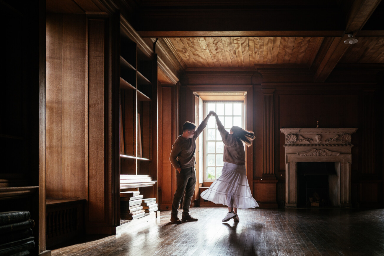 A Secret Proposal at Howth Cliffs & a Private Hour at Howth Castle: Joseph & Shannon's Engagement Shoot 18 Two people dance in an empty wood-panelled room with large windows and a fireplace, sunlight streaming in and highlighting their movement.