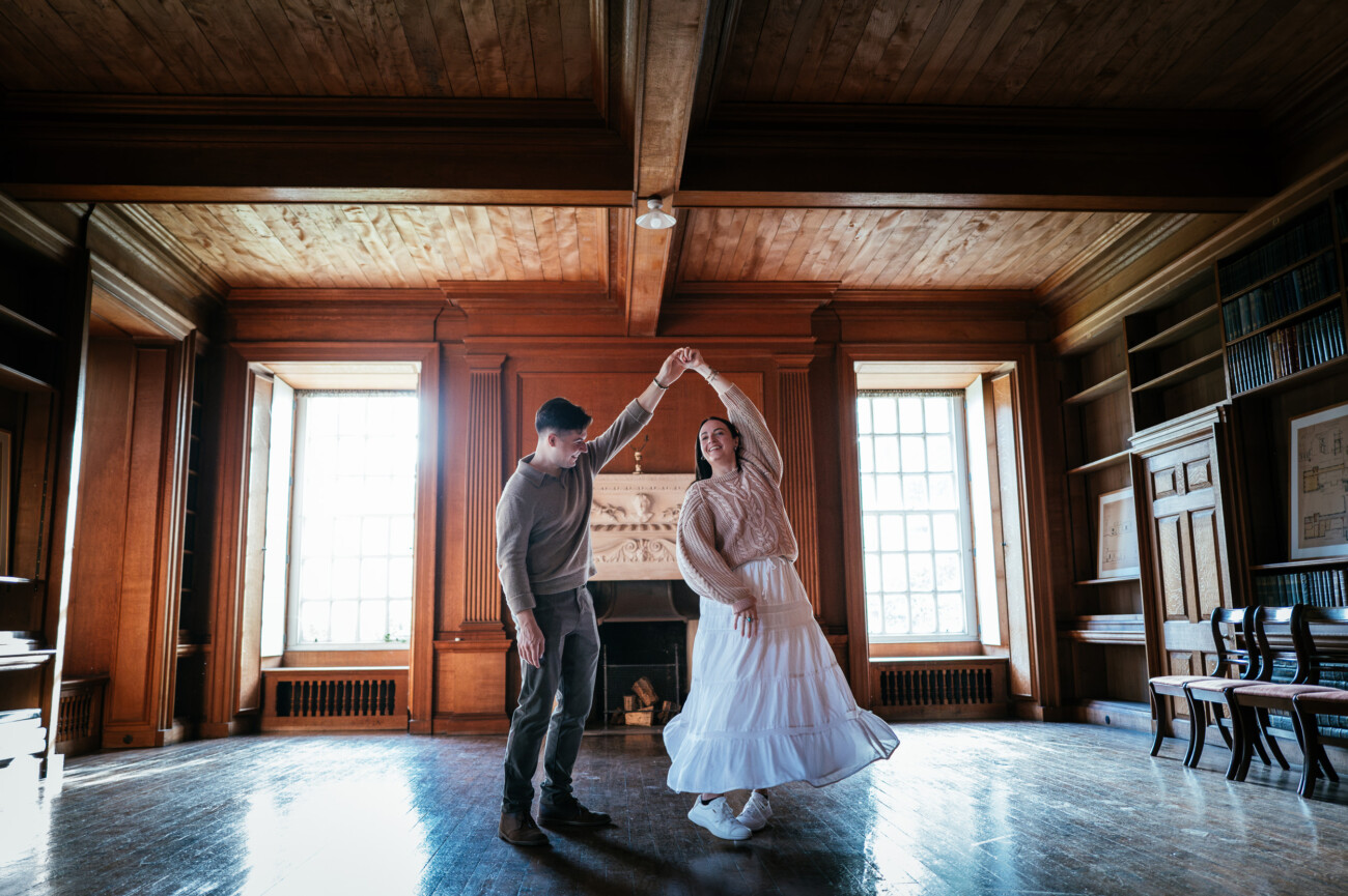 A Secret Proposal at Howth Cliffs & a Private Hour at Howth Castle: Joseph & Shannon's Engagement Shoot 19 Two people dance together in a spacious wooden room with large windows, bookshelves, and a fireplace.