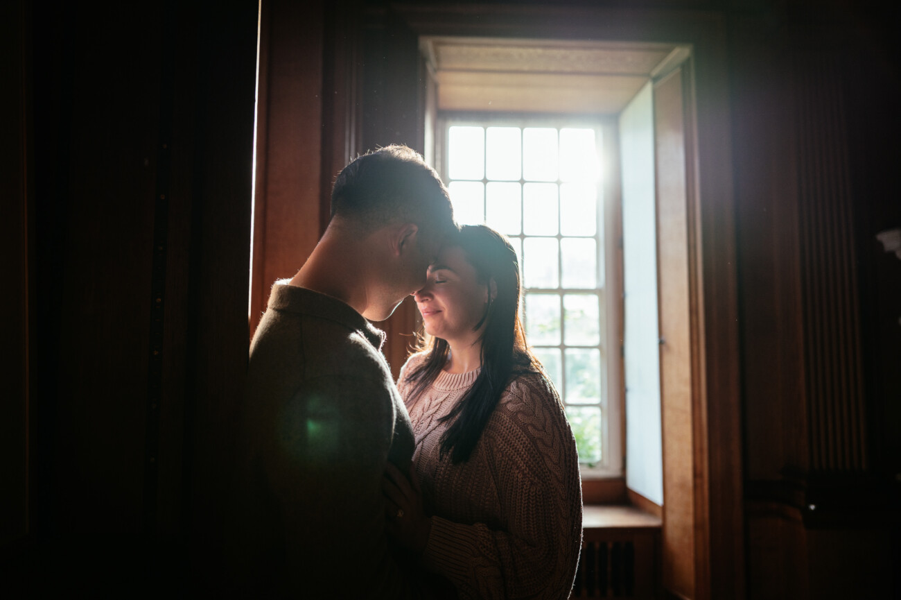 A Secret Proposal at Howth Cliffs & a Private Hour at Howth Castle: Joseph & Shannon's Engagement Shoot 20 A couple stands closely together by a sunlit window in a dimly lit room, facing each other with gentle expressions.