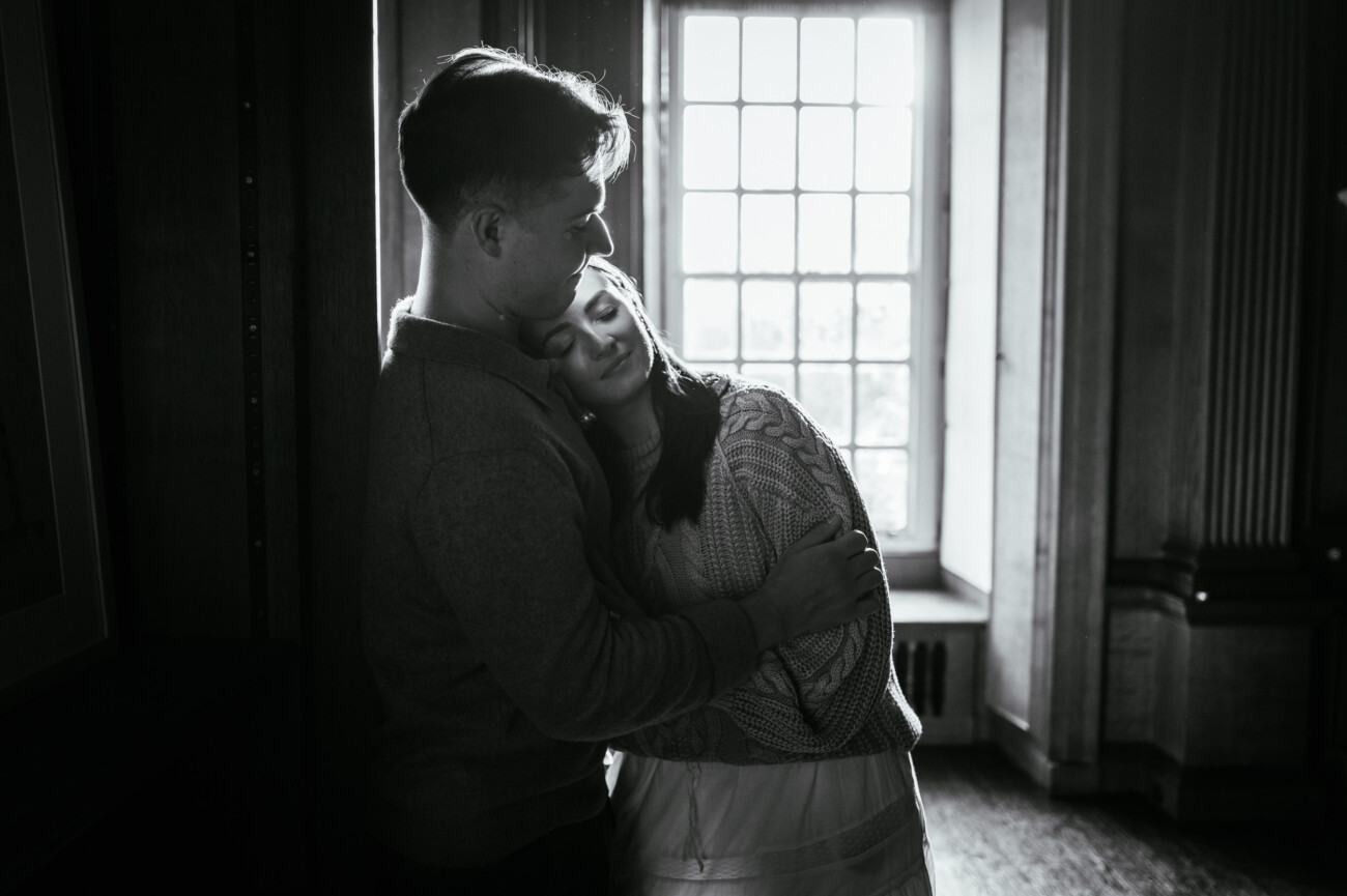 A Secret Proposal at Howth Cliffs & a Private Hour at Howth Castle: Joseph & Shannon's Engagement Shoot 21 A couple stands in front of a window, embracing closely; the woman rests her head on the man’s chest with her eyes closed. The image is in black and white.