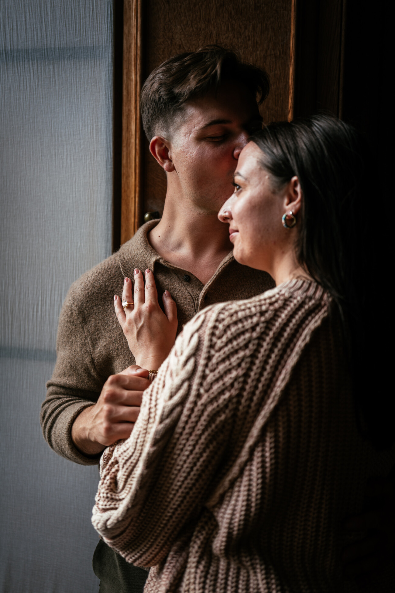 A Secret Proposal at Howth Cliffs & a Private Hour at Howth Castle: Joseph & Shannon's Engagement Shoot 22 A man kisses a woman’s forehead as she rests her hand on his chest, both wearing knitted jumpers and standing near a window.