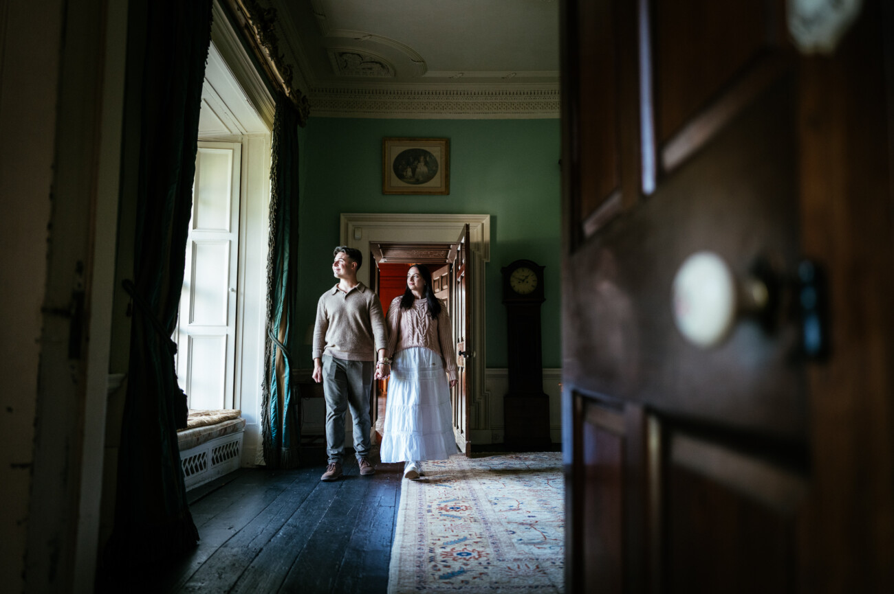 A Secret Proposal at Howth Cliffs & a Private Hour at Howth Castle: Joseph & Shannon's Engagement Shoot 23 A man and woman in vintage-style clothing stand side by side in a corridor of an old, elegant house with green walls and large windows.