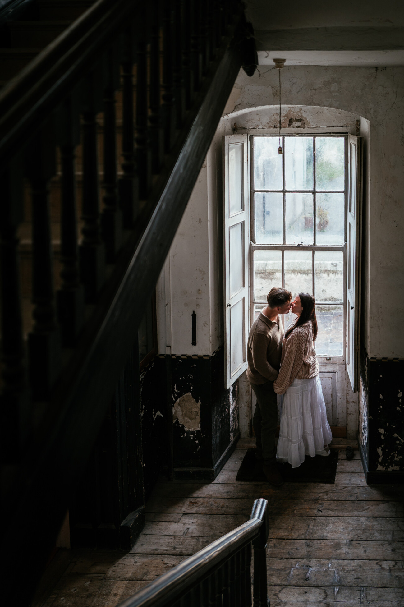 A Secret Proposal at Howth Cliffs & a Private Hour at Howth Castle: Joseph & Shannon's Engagement Shoot 24 Two people stand close together near a tall window in an old, worn interior with wooden floors and a staircase visible in the foreground.
