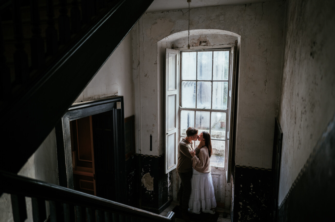A Secret Proposal at Howth Cliffs & a Private Hour at Howth Castle: Joseph & Shannon's Engagement Shoot 25 A couple stands close together by a tall window in an old, dimly lit stairwell with peeling walls and natural light streaming in at Howth Castle Dublin.