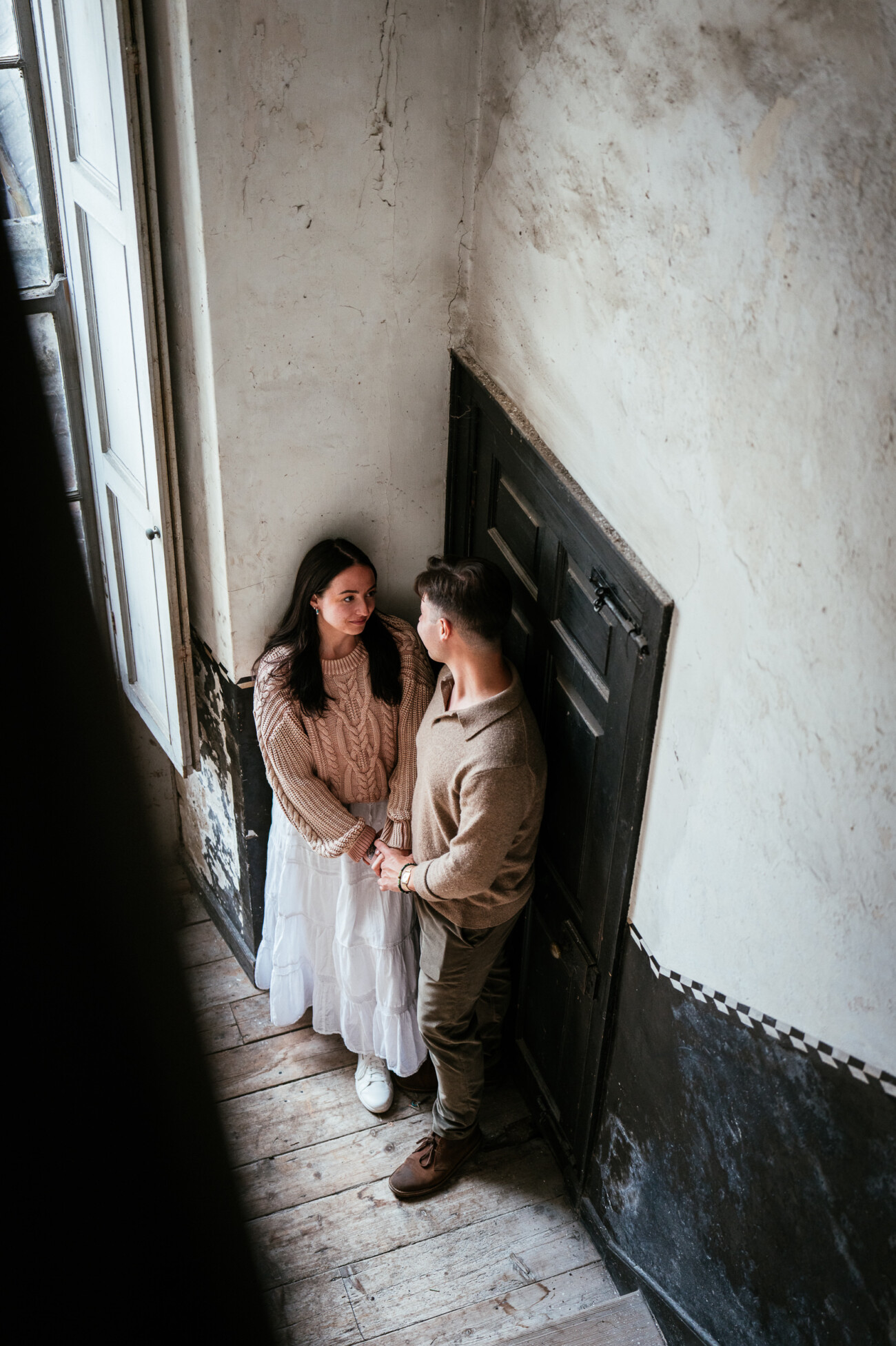 A Secret Proposal at Howth Cliffs & a Private Hour at Howth Castle: Joseph & Shannon's Engagement Shoot 26 A woman and man stand close together in a dimly lit corner near a window, holding hands and looking at each other.