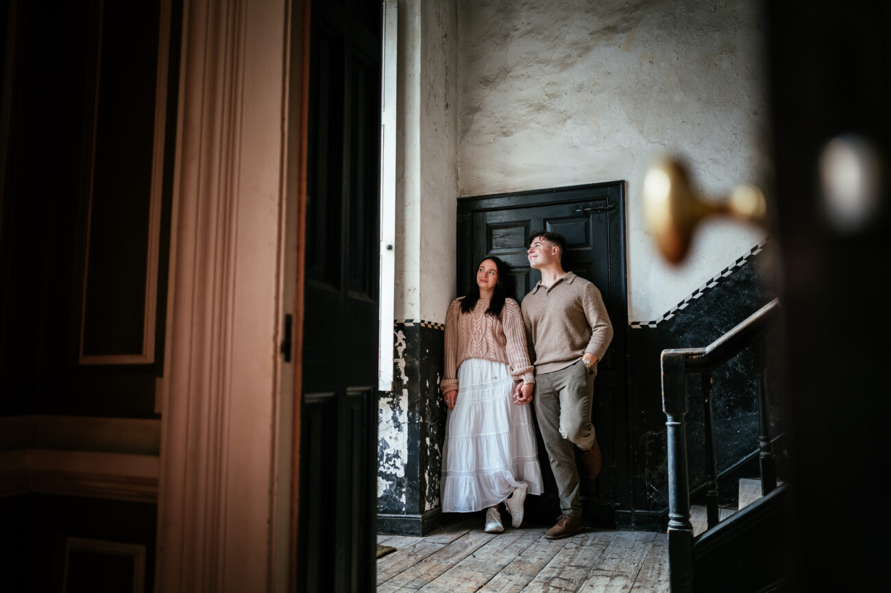 A Secret Proposal at Howth Cliffs & a Private Hour at Howth Castle: Joseph & Shannon's Engagement Shoot 27 A couple stands holding hands in an old, dimly lit building near a staircase, looking up towards the light from a window.