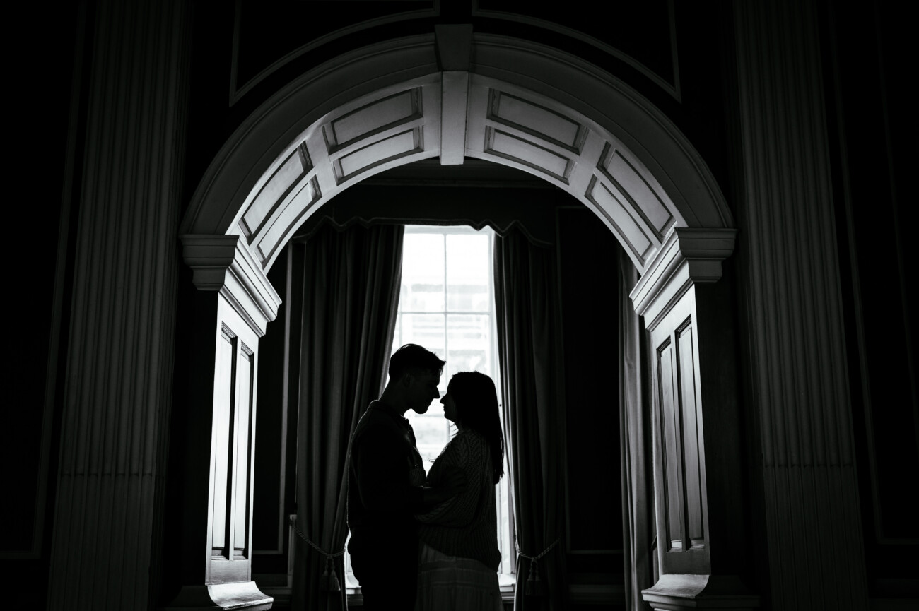 A Secret Proposal at Howth Cliffs & a Private Hour at Howth Castle: Joseph & Shannon's Engagement Shoot 28 Two people stand close together in silhouette beneath an ornate archway, with light streaming through a window behind them.