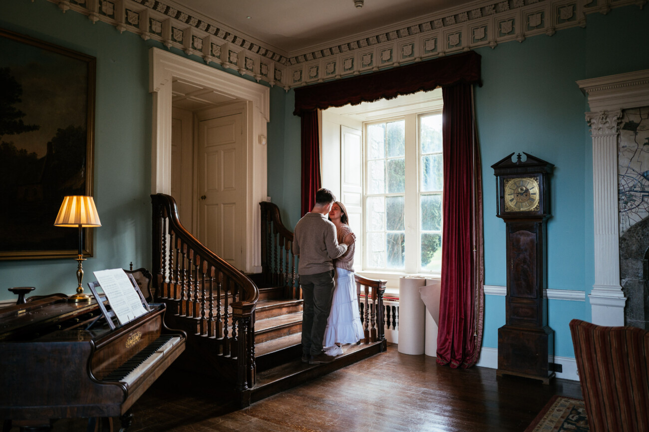 A Secret Proposal at Howth Cliffs & a Private Hour at Howth Castle: Joseph & Shannon's Engagement Shoot 29 A couple stands together at the bottom of a wooden staircase near a large window in a traditionally decorated room with a piano and grandfather clock.