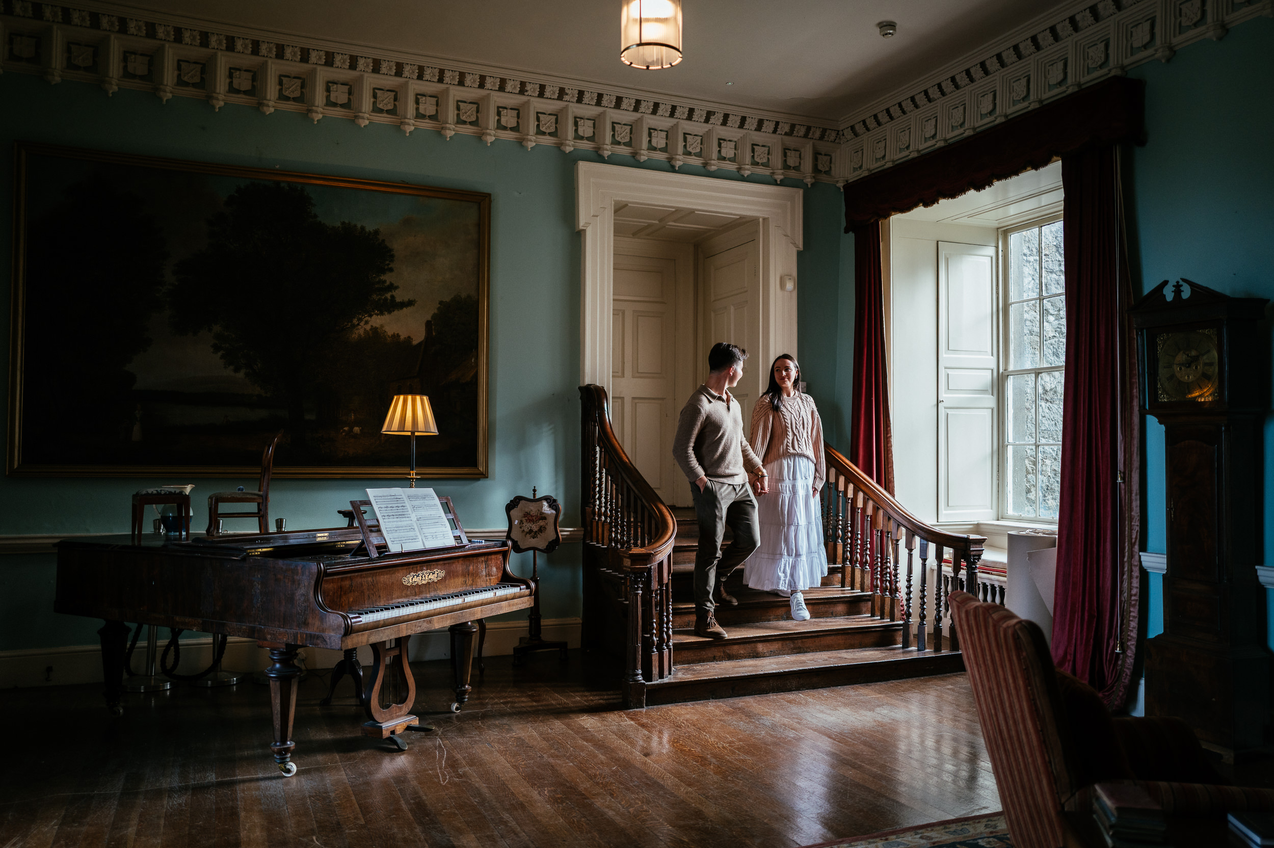 An engaged couple stand on a wooden stairway in a room with a grand piano, large painting, and window with red curtains during their Howth Castle private engagement photoshoot