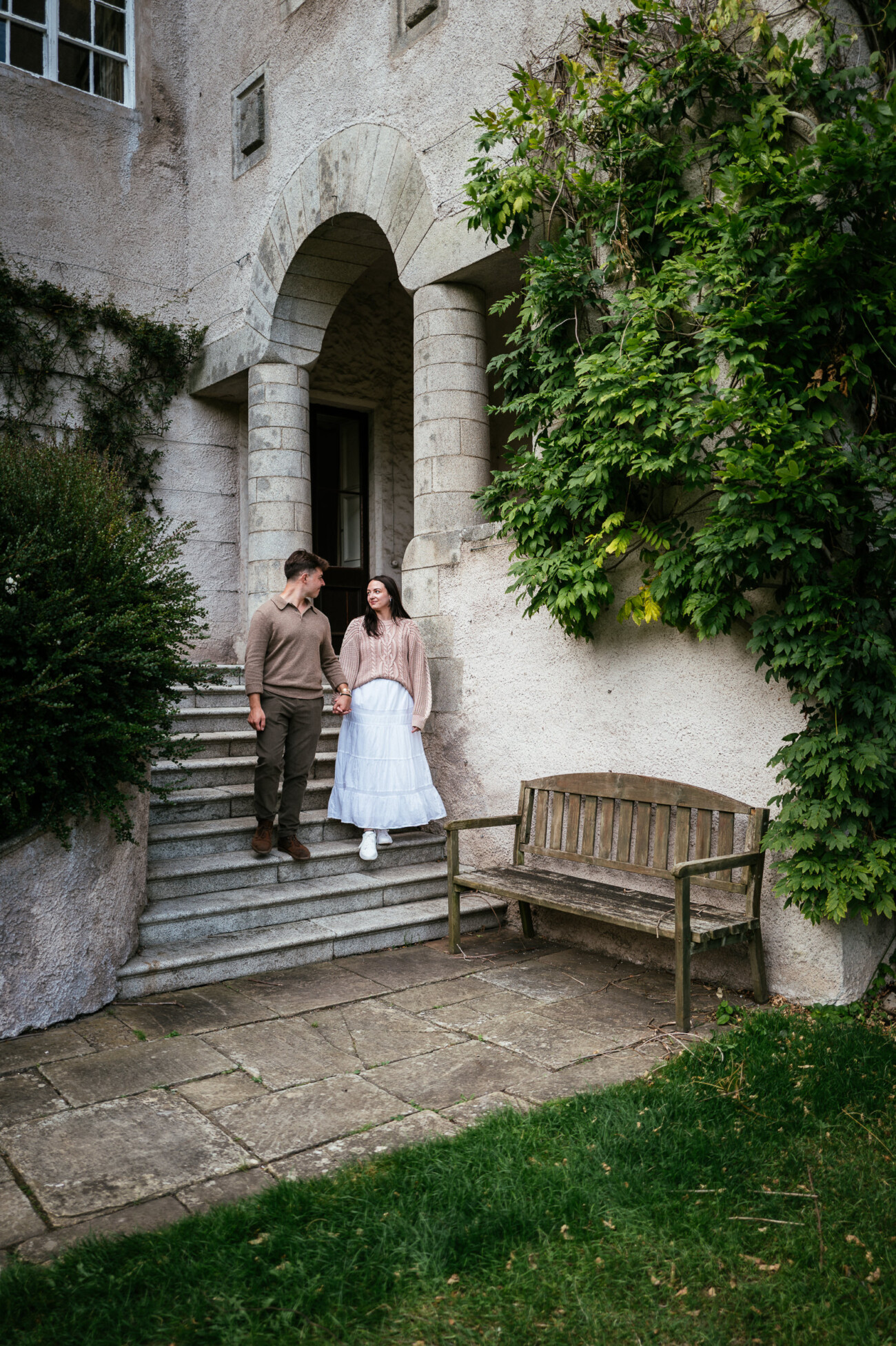 A Secret Proposal at Howth Cliffs & a Private Hour at Howth Castle: Joseph & Shannon's Engagement Shoot 32 A man and a woman hold hands whilst standing on stone steps outside a building covered in greenery, with a wooden bench nearby.