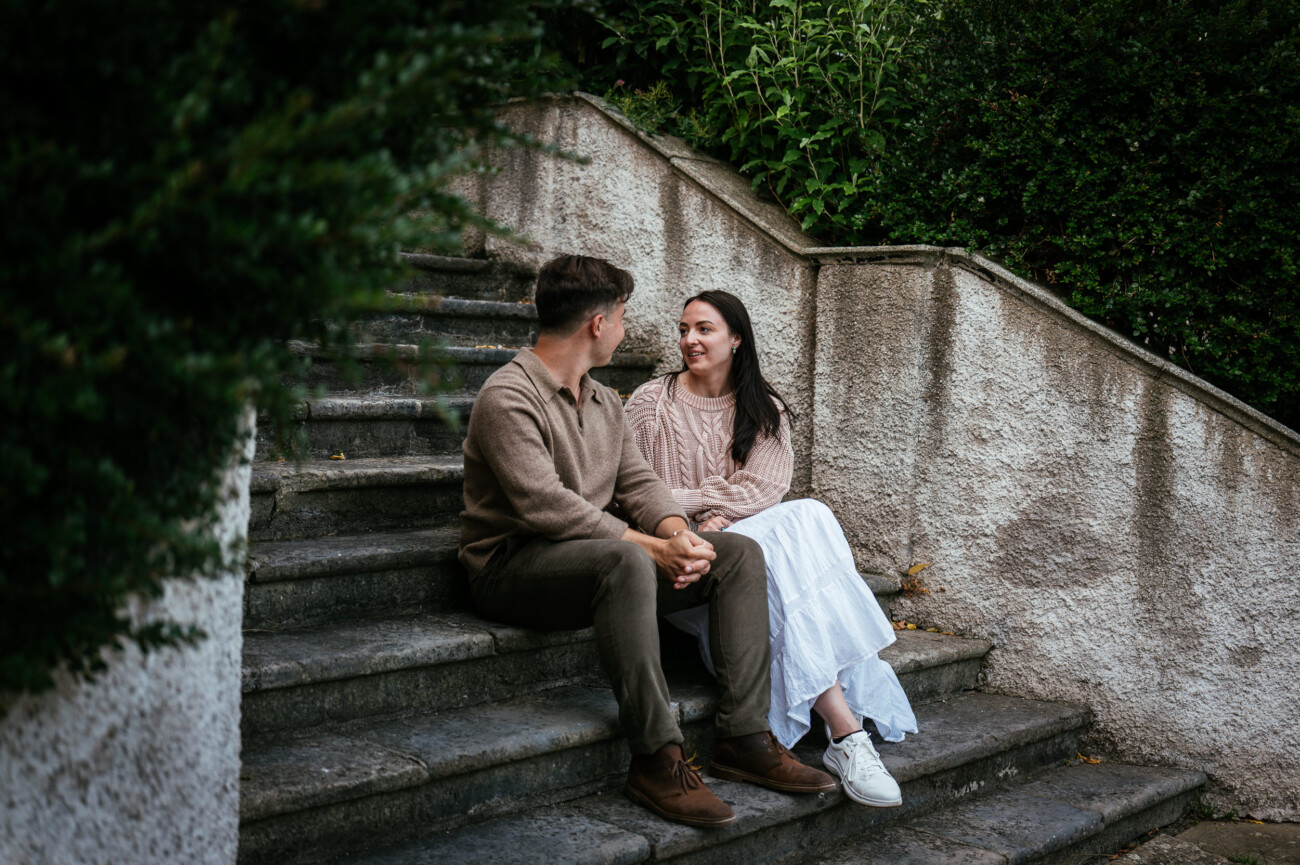 A Secret Proposal at Howth Cliffs & a Private Hour at Howth Castle: Joseph & Shannon's Engagement Shoot 33 A man and a woman sit on outdoor stone steps, holding hands and smiling at each other, surrounded by greenery.
