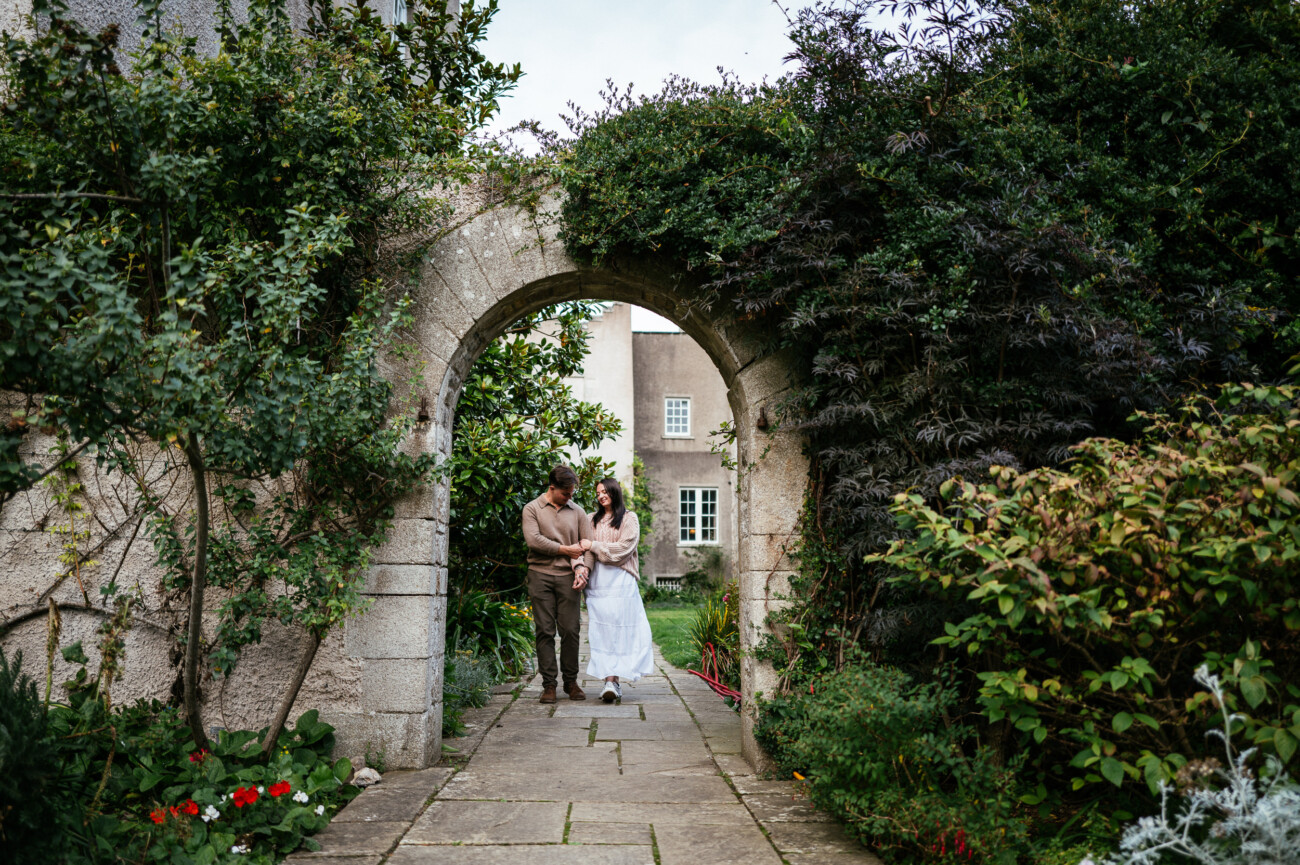 A Secret Proposal at Howth Cliffs & a Private Hour at Howth Castle: Joseph & Shannon's Engagement Shoot 34 A couple stands under a stone archway in a lush garden, holding hands and looking at each other, with a building visible in the background.