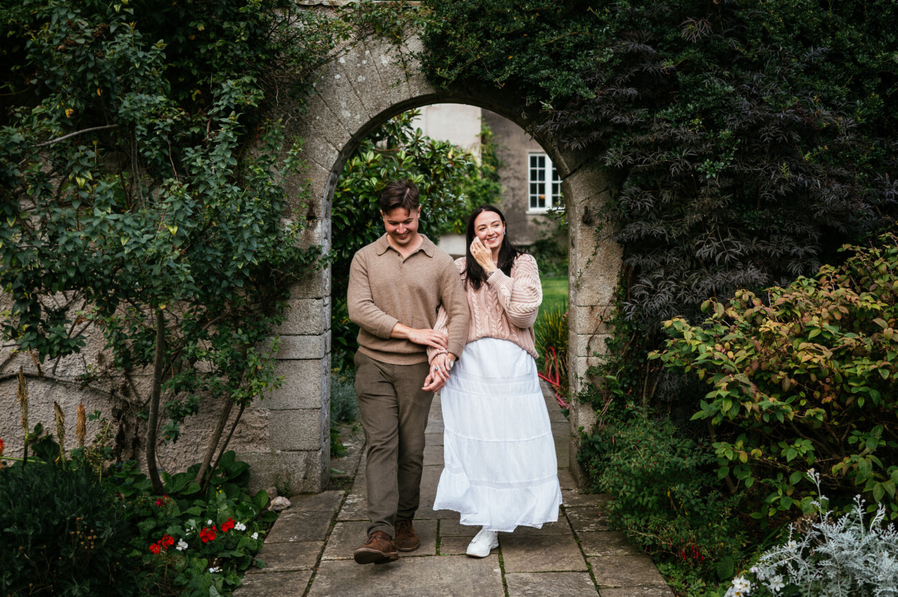 A Secret Proposal at Howth Cliffs & a Private Hour at Howth Castle: Joseph & Shannon's Engagement Shoot 35 A man and woman walk arm in arm through a stone archway surrounded by greenery and plants, smiling at each other.