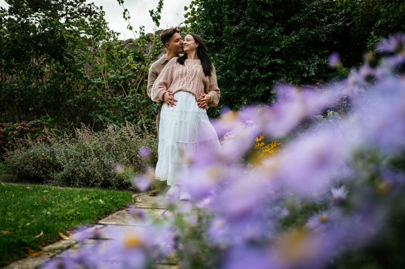A Secret Proposal at Howth Cliffs & a Private Hour at Howth Castle: Joseph & Shannon's Engagement Shoot 36 A couple stand together on a garden path, with the man embracing the woman from behind. Purple flowers are blurred in the foreground, and green foliage surrounds them.