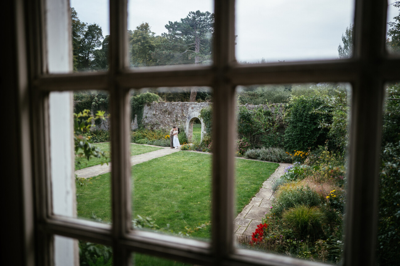 A Secret Proposal at Howth Cliffs & a Private Hour at Howth Castle: Joseph & Shannon's Engagement Shoot 37 A couple stands together in a garden courtyard, viewed through a window with multiple panes. Greenery and flowers surround the stone path and lawn.