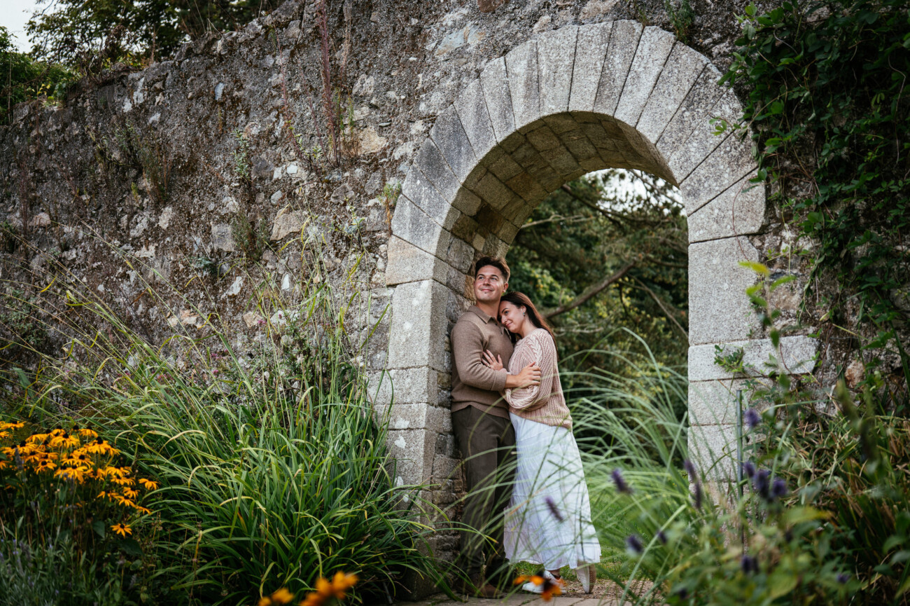 A Secret Proposal at Howth Cliffs & a Private Hour at Howth Castle: Joseph & Shannon's Engagement Shoot 38 A couple stands closely together under a stone archway surrounded by greenery and flowers in a garden setting.