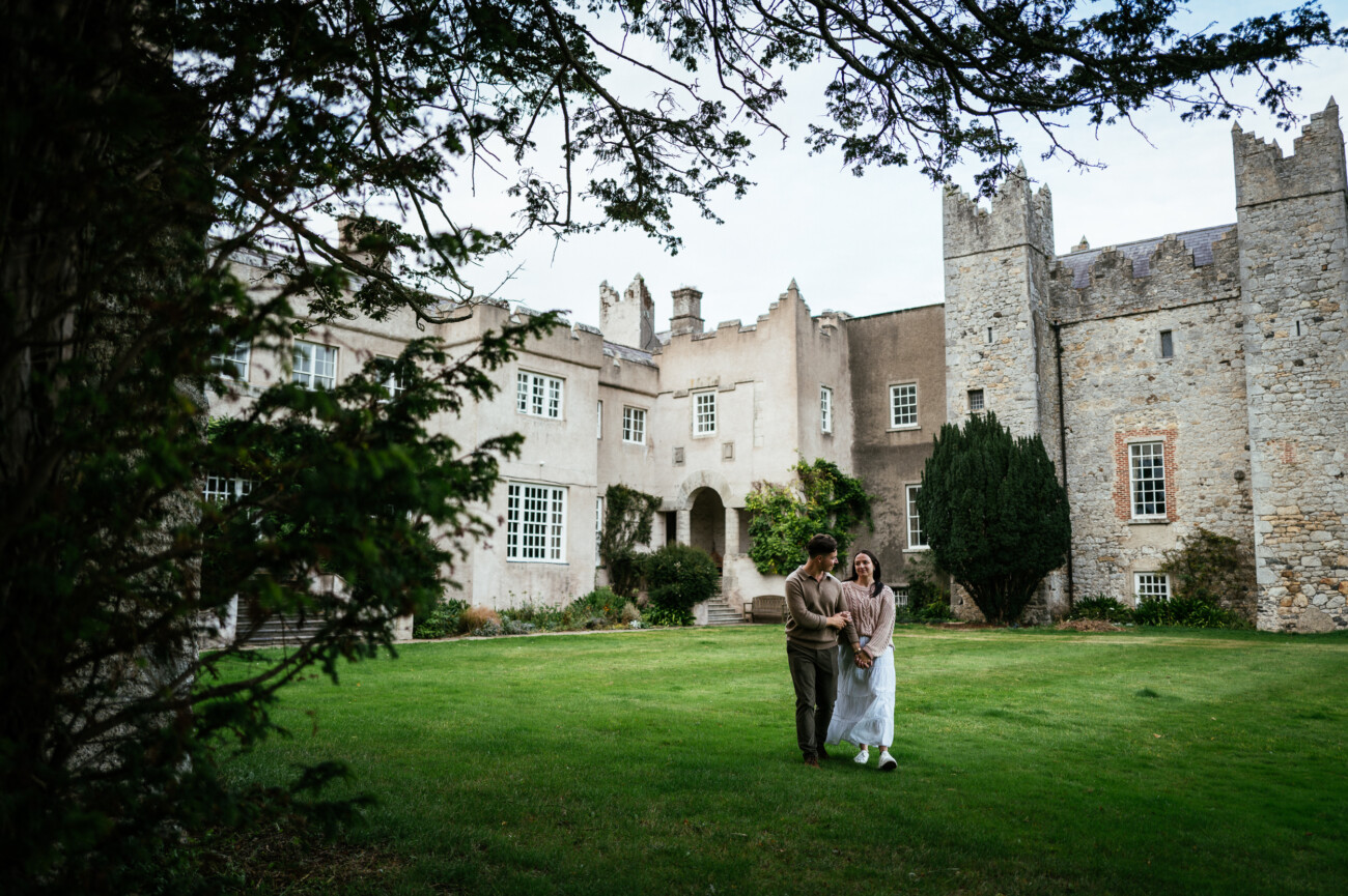 A Secret Proposal at Howth Cliffs & a Private Hour at Howth Castle: Joseph & Shannon's Engagement Shoot 39 A couple walks arm in arm on the lawn in front of a large stone building with castle-like features and greenery.