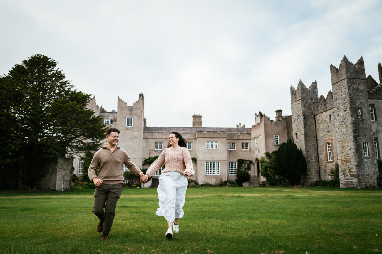 A Secret Proposal at Howth Cliffs & a Private Hour at Howth Castle: Joseph & Shannon's Engagement Shoot 40 A man and woman holding hands run across the grass in front of a large stone castle on a cloudy day.