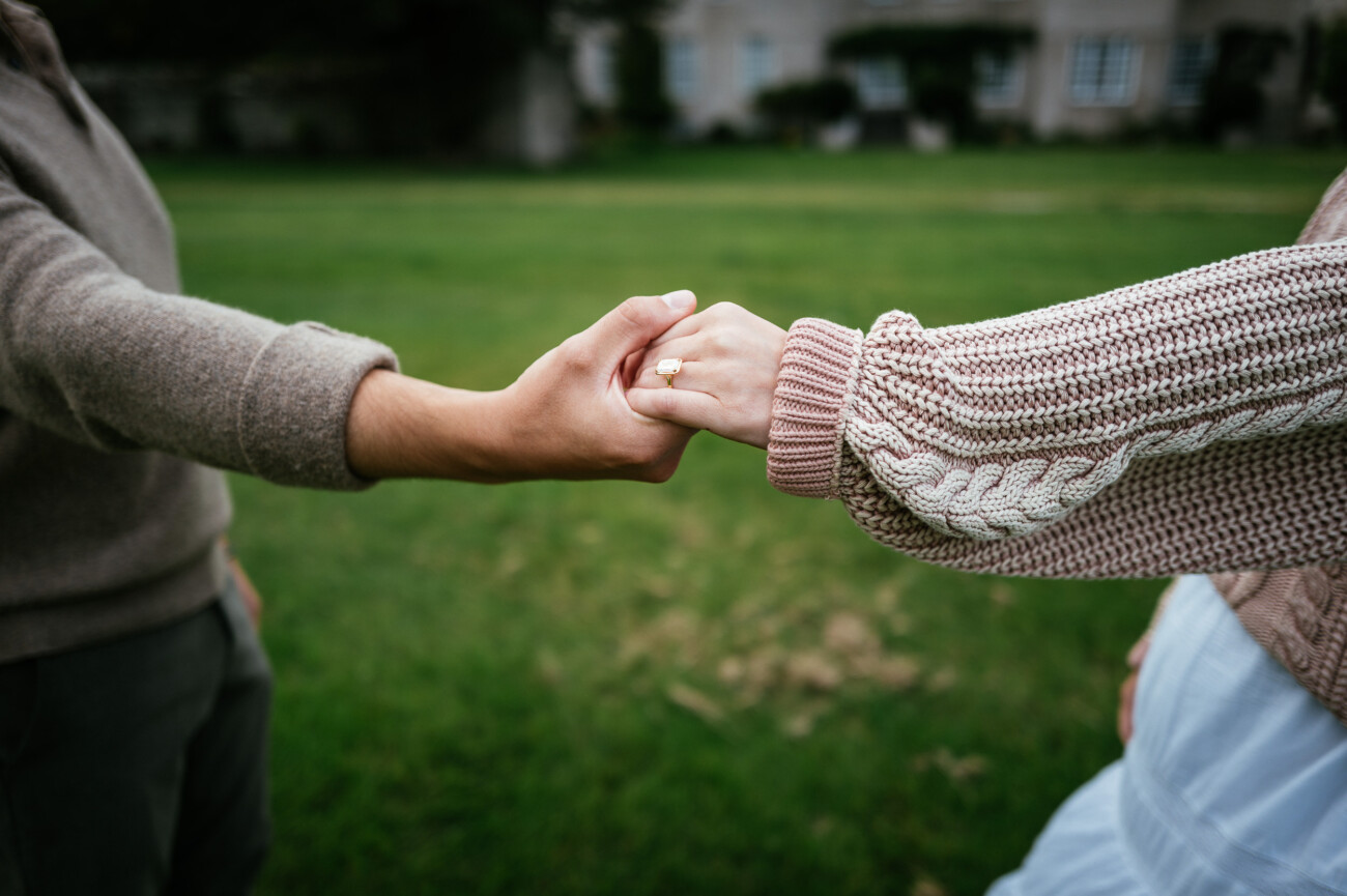 A Secret Proposal at Howth Cliffs & a Private Hour at Howth Castle: Joseph & Shannon's Engagement Shoot 41 Two people are holding hands outdoors. One person is wearing a ring and a knitted jumper; the background is a grassy area with a building visible.