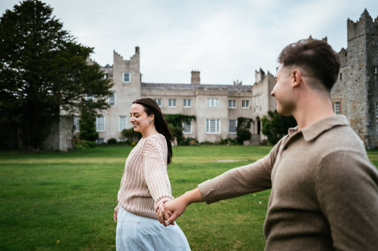 A Secret Proposal at Howth Cliffs & a Private Hour at Howth Castle: Joseph & Shannon's Engagement Shoot 42 A woman and a man hold hands and walk on grass in front of a large stone building with multiple windows and towers, under a cloudy sky.