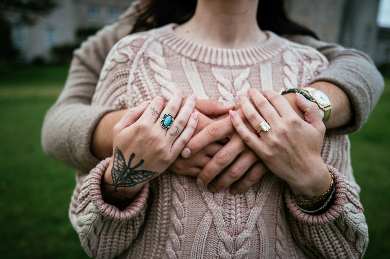 A Secret Proposal at Howth Cliffs & a Private Hour at Howth Castle: Joseph & Shannon's Engagement Shoot 43 A person stands behind another, hugging them with both arms. The front person wears a knitted jumper and rings, showing a butterfly hand tattoo. Both pairs of hands are visible.