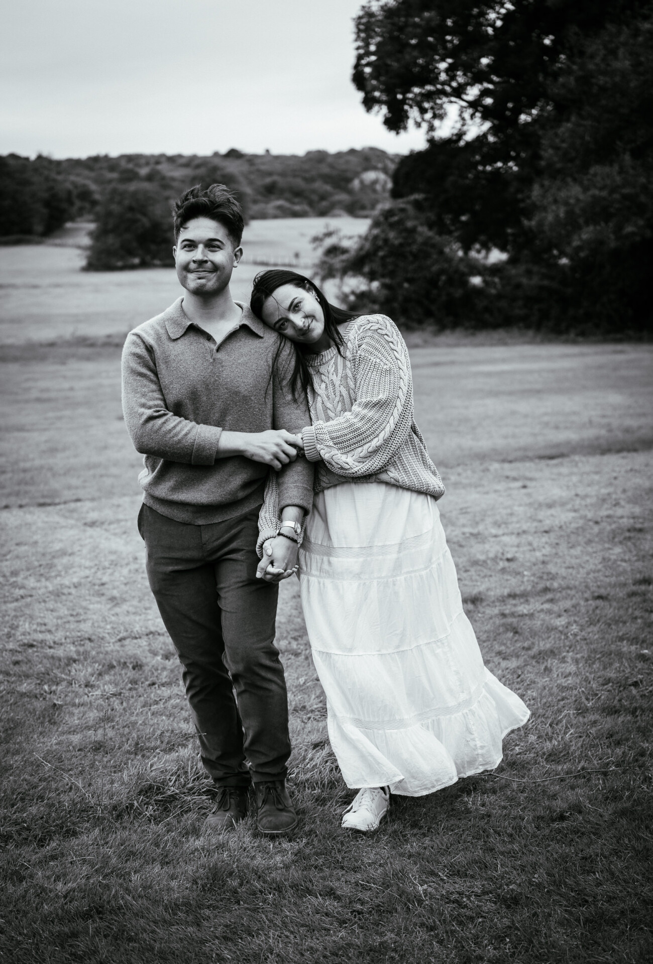A Secret Proposal at Howth Cliffs & a Private Hour at Howth Castle: Joseph & Shannon's Engagement Shoot 44 A man and woman stand on grass in a field. The woman leans her head on the man’s shoulder and they hold hands. Both wear casual clothes. Trees and open land are in the background.