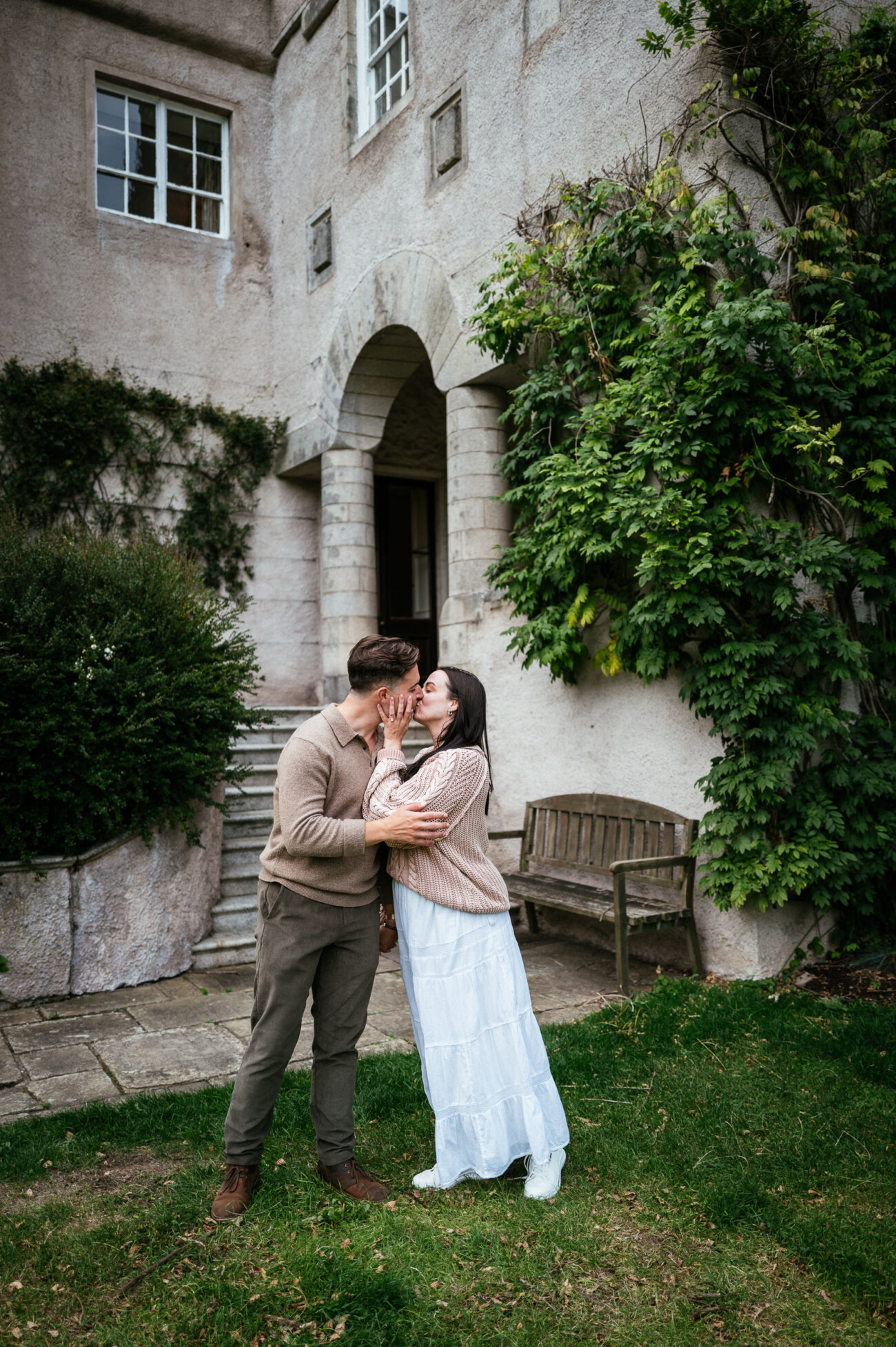 A Secret Proposal at Howth Cliffs & a Private Hour at Howth Castle: Joseph & Shannon's Engagement Shoot 45 A couple stands outside a stone building, kissing. The woman wears a white skirt and beige jumper; the man wears olive trousers and a tan jumper. Greenery surrounds the entrance behind them.