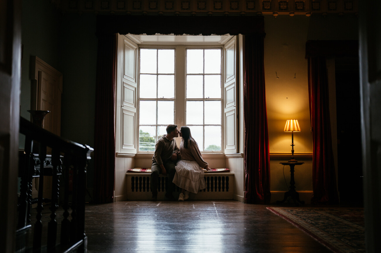 A Secret Proposal at Howth Cliffs & a Private Hour at Howth Castle: Joseph & Shannon's Engagement Shoot 46 A couple sits closely on a window seat, facing a large window with white frames. Soft daylight filters in, and a lit lamp stands to the right.