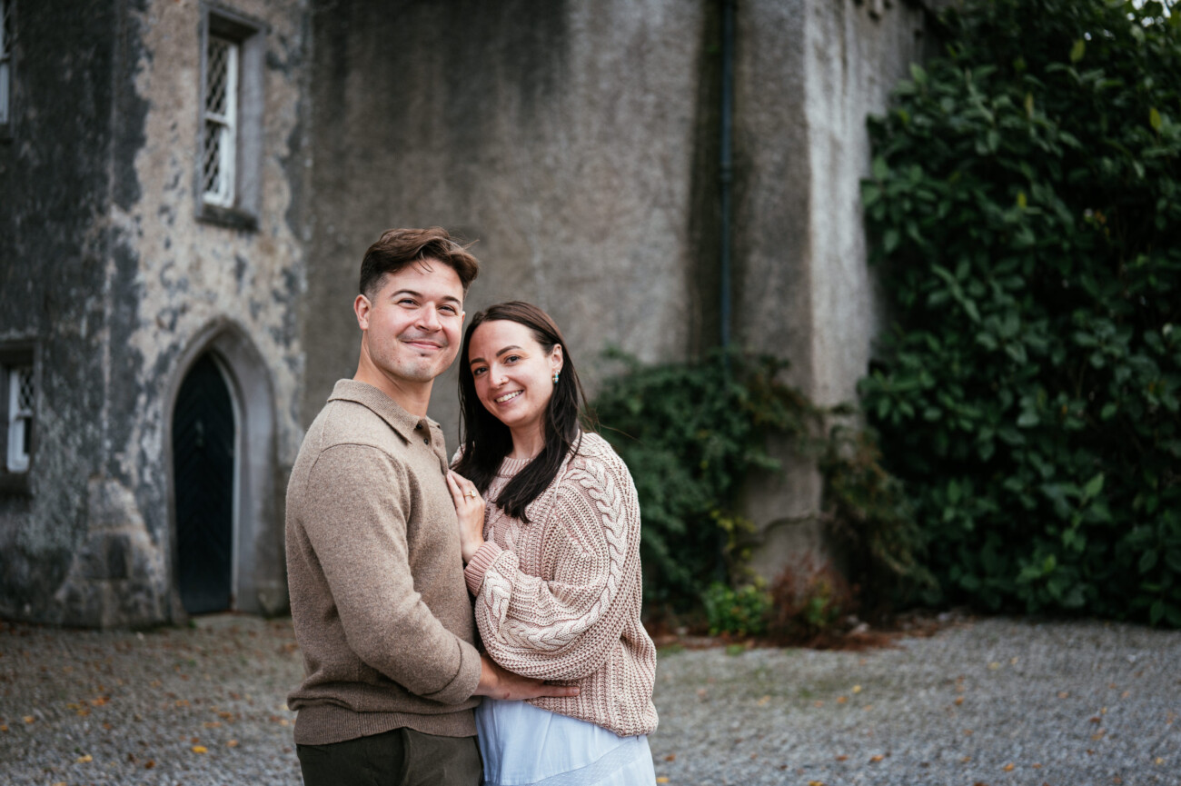 A Secret Proposal at Howth Cliffs & a Private Hour at Howth Castle: Joseph & Shannon's Engagement Shoot 47 A couple stands close together, smiling at the camera in front of an old stone building with greenery and a gravel ground.