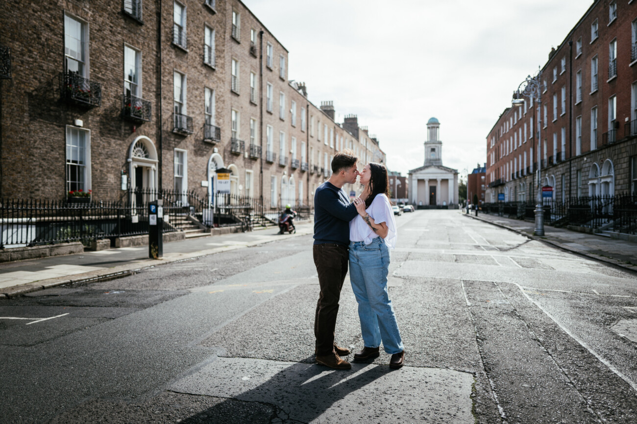 A Secret Proposal at Howth Cliffs & a Private Hour at Howth Castle: Joseph & Shannon's Engagement Shoot 48 A couple stands close together in the middle of an empty street lined with brick buildings, with a church visible in the distance under a cloudy sky.