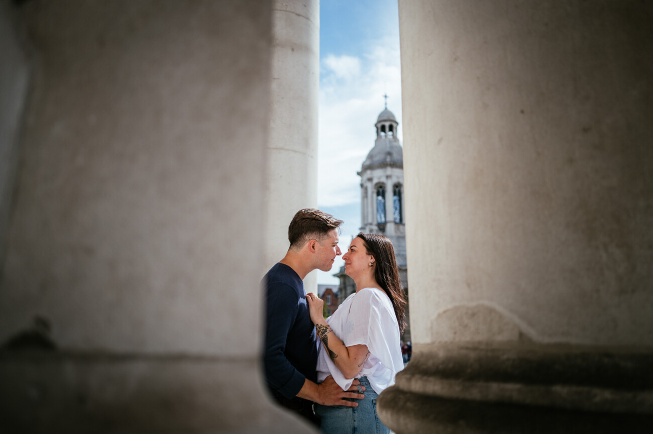 A Secret Proposal at Howth Cliffs & a Private Hour at Howth Castle: Joseph & Shannon's Engagement Shoot 58 A man and woman stand close together between large stone columns, with a domed building in the background under a blue sky.