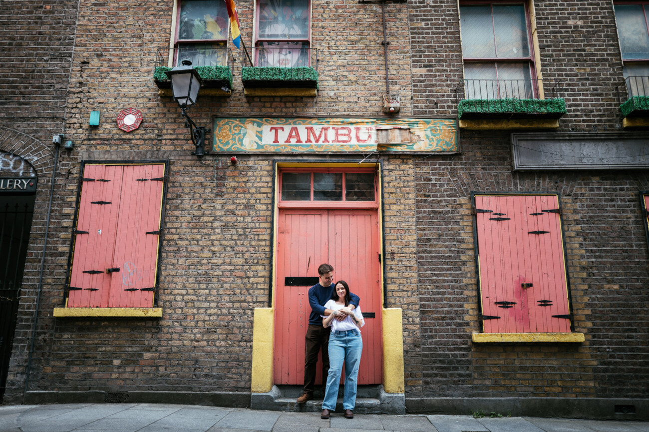 A Secret Proposal at Howth Cliffs & a Private Hour at Howth Castle: Joseph & Shannon's Engagement Shoot 59 A man and woman stand in front of a brick building with pink shutters and a sign that reads “TAMBURY.” The woman holds a small dog, and both look at the camera.