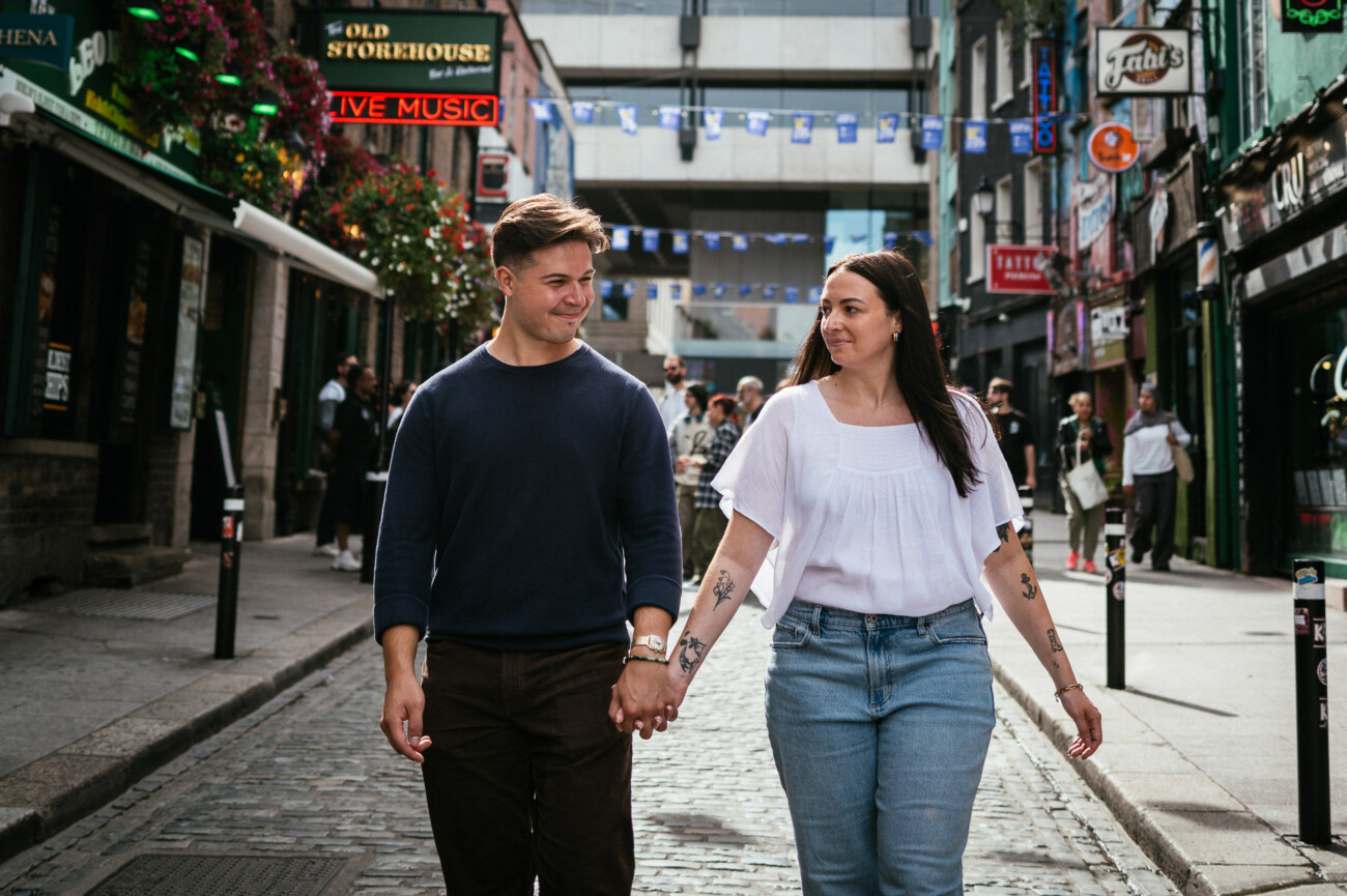 A Secret Proposal at Howth Cliffs & a Private Hour at Howth Castle: Joseph & Shannon's Engagement Shoot 60 A man and woman hold hands while walking down a cobbled street in a lively city area with shops and people in the background.