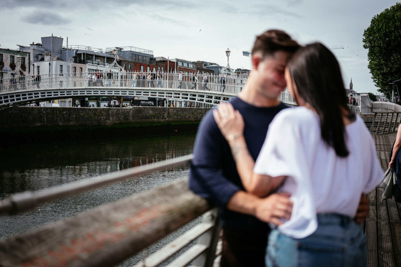 A Secret Proposal at Howth Cliffs & a Private Hour at Howth Castle: Joseph & Shannon's Engagement Shoot 61 A couple stands close together on a riverside walkway, with a white footbridge and city buildings in the background.