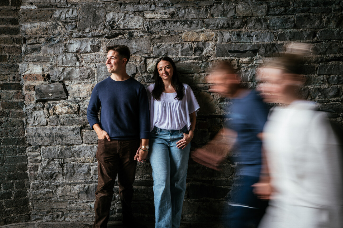 A Secret Proposal at Howth Cliffs & a Private Hour at Howth Castle: Joseph & Shannon's Engagement Shoot 62 Two people stand holding hands against a stone wall whilst two blurred figures walk past in the foreground.